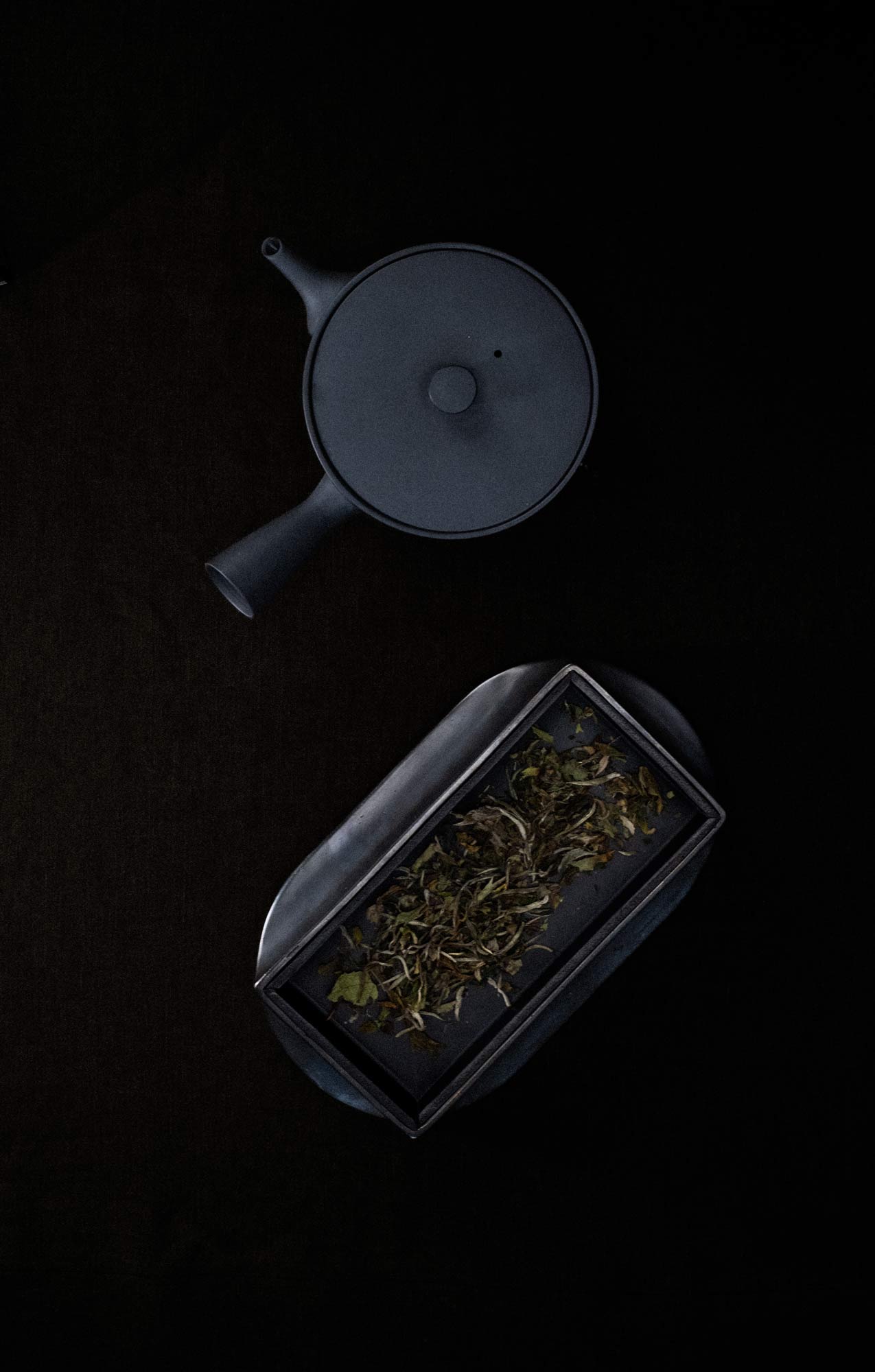 Optimierter Alt-Text:
Kyusu von oben fotografiert auf einem Tisch mit schwarzem Tischtuch, daneben ein gusseiserner, länglicher Teller mit hohen Wänden, der Teeblätter enthält.